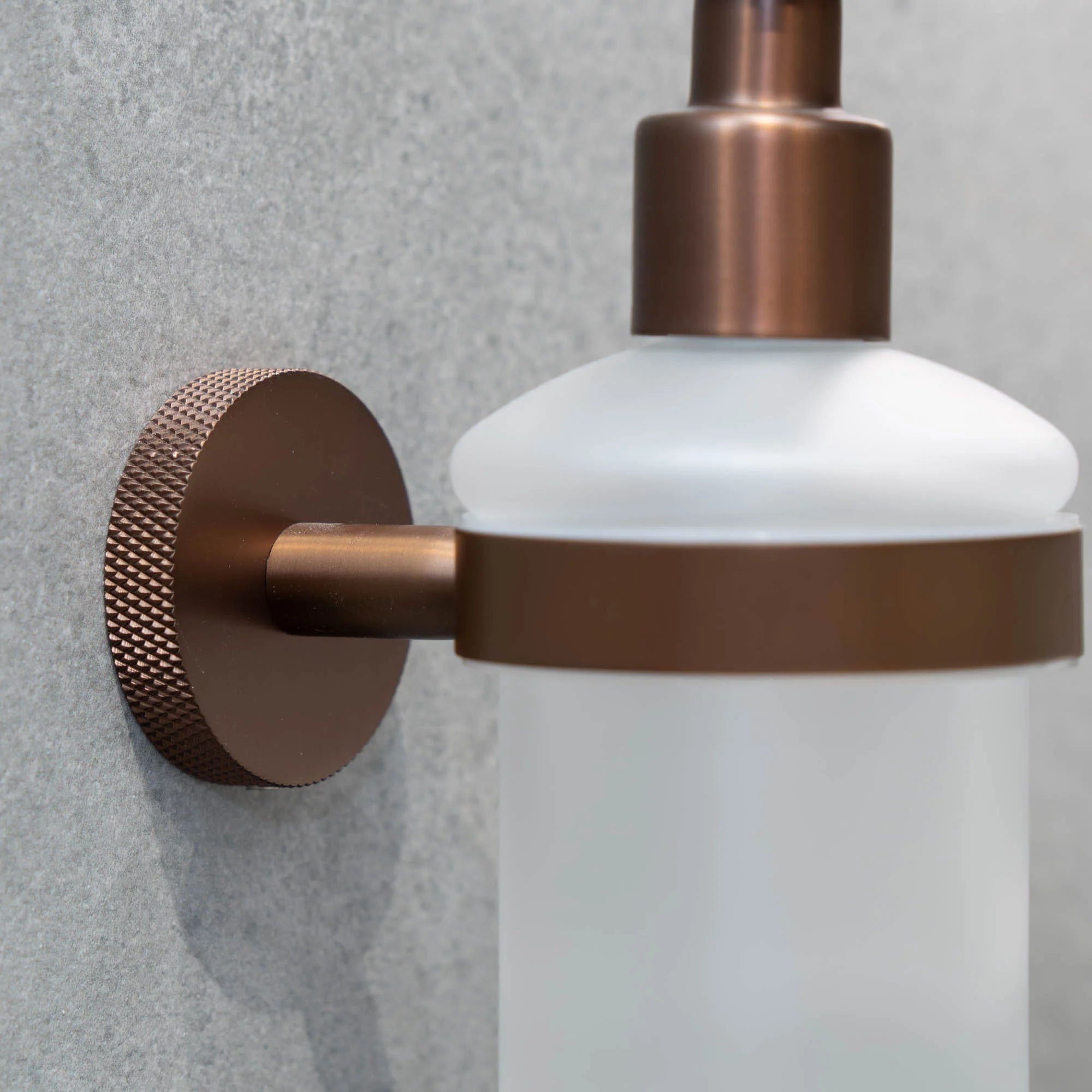 Wall-mounted portofino soap dispenser with bronze pump and frosted container on a grey wall deluxe bathrooms